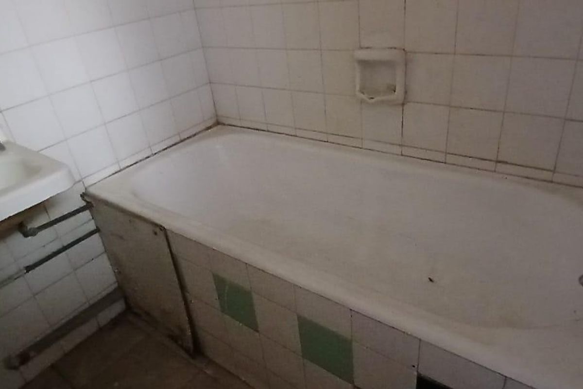 A dated bathroom featuring a standard bathtub and wall-mounted basin with exposed plumbing and worn ceramic tiling throughout.