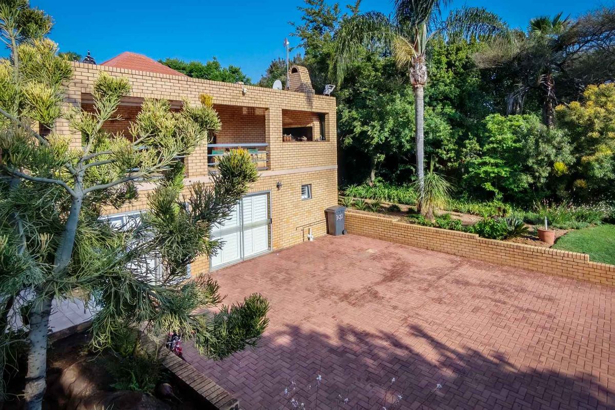 Rear view of a face brick house featuring a paved driveway, a balcony with a built-in braai, and a landscaped garden area.