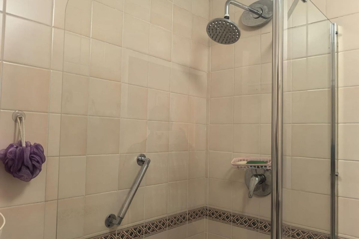 A functional shower enclosure featuring cream wall tiles with a decorative border, a fixed shower head, and a safety grab rail.