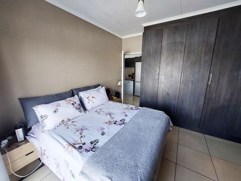 A bedroom featuring dark wood built-in cupboards and tiled flooring, with a view into the adjacent kitchen area.