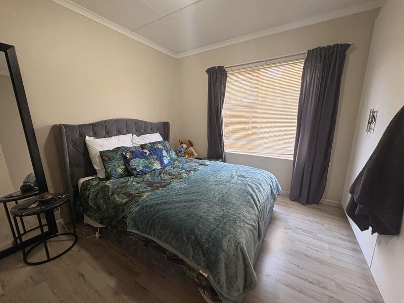 A well-maintained bedroom featuring light-toned laminate flooring, neutral wall finishes, and window treatments including venetian blinds and curtains.