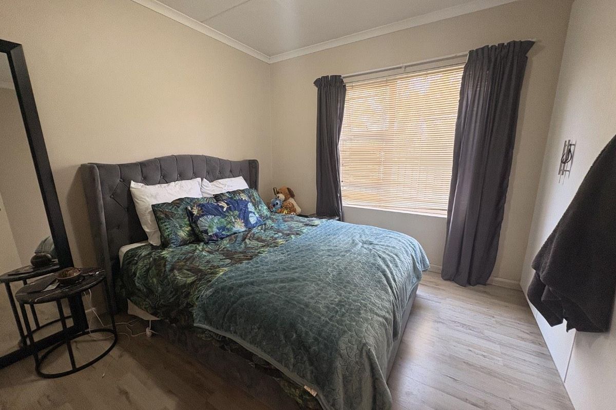A well-maintained bedroom featuring light-toned laminate flooring, neutral wall finishes, and window treatments including venetian blinds and curtains.