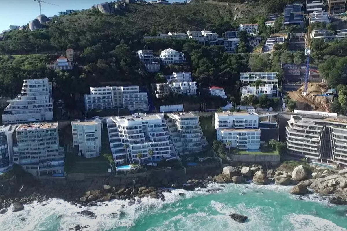An aerial view of a coastal apartment complex nestled against a mountain slope, showcasing the building's proximity to the ocean.