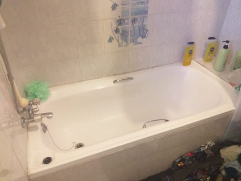 A functional bathroom featuring a built-in bathtub with chrome fixtures and decorative wall tiling, requiring minor maintenance to grout and sealant.