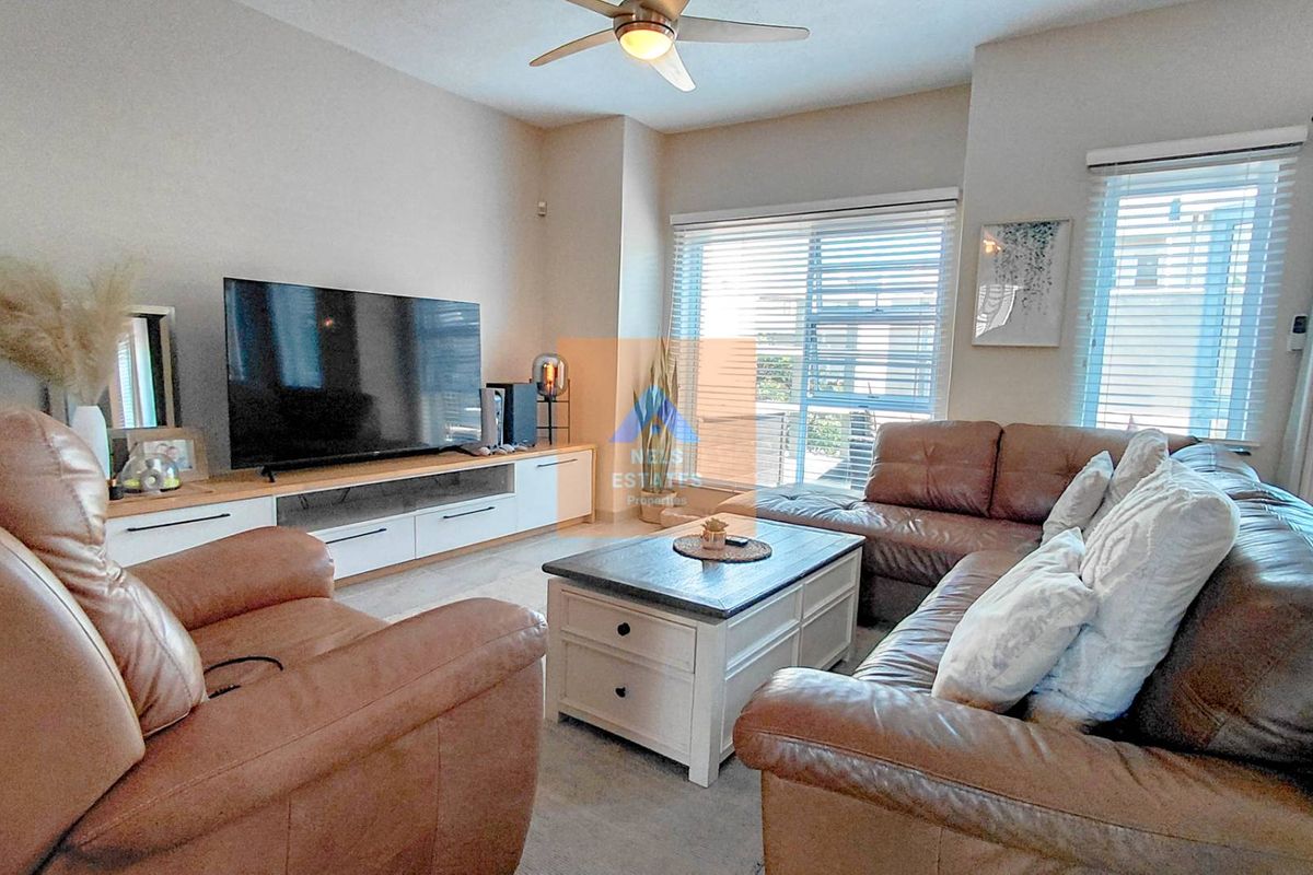 A comfortable living room featuring a ceiling fan, large windows with venetian blinds, and access to an outdoor area via a sliding glass door.
