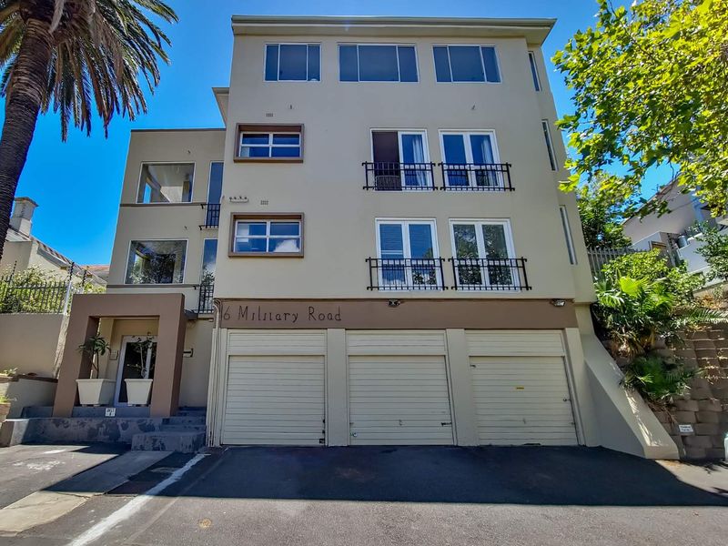 Front elevation of a multi-story apartment building featuring three garage doors, balconies with railings, and a paved driveway entrance.