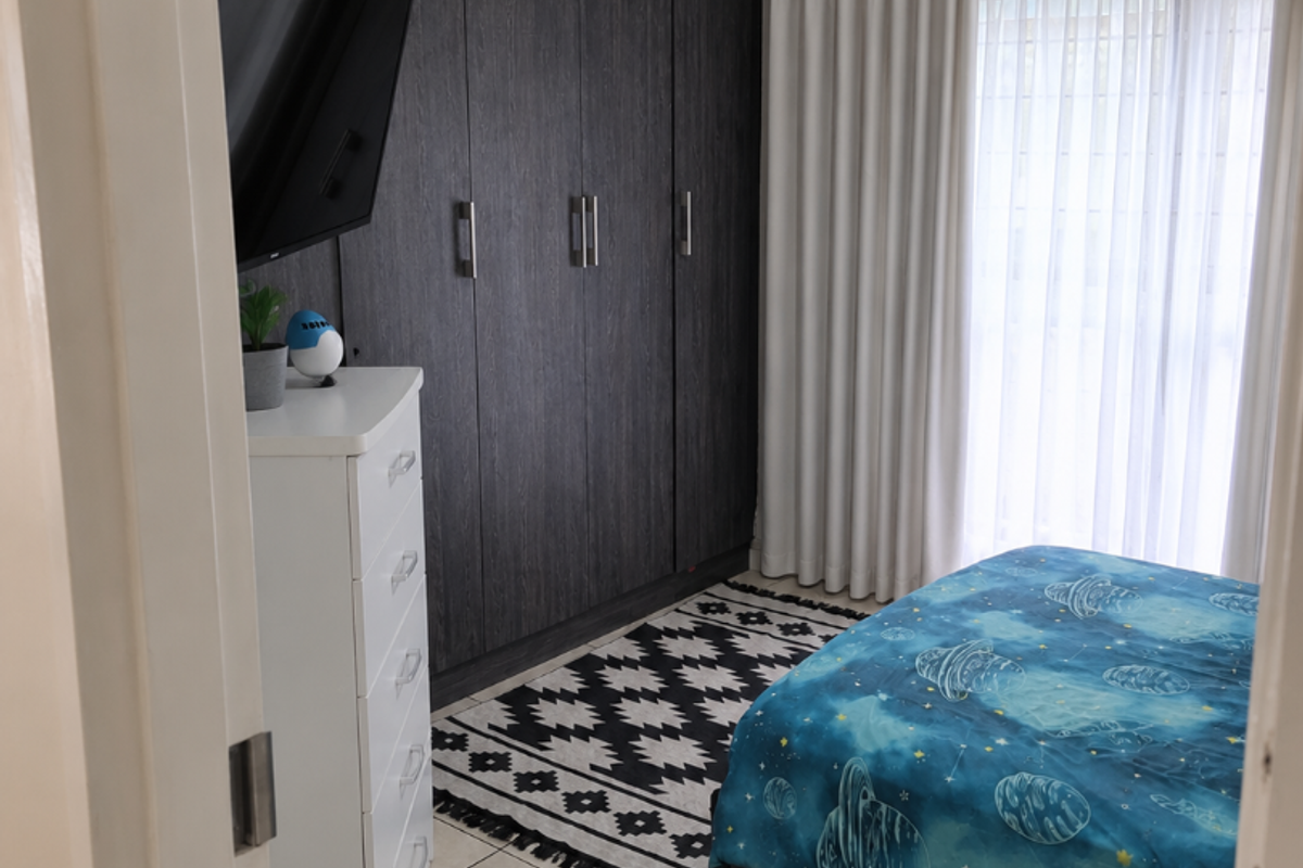 A well-maintained bedroom featuring dark wood built-in cupboards, neutral tiled flooring, and large windows with floor-to-ceiling curtains.