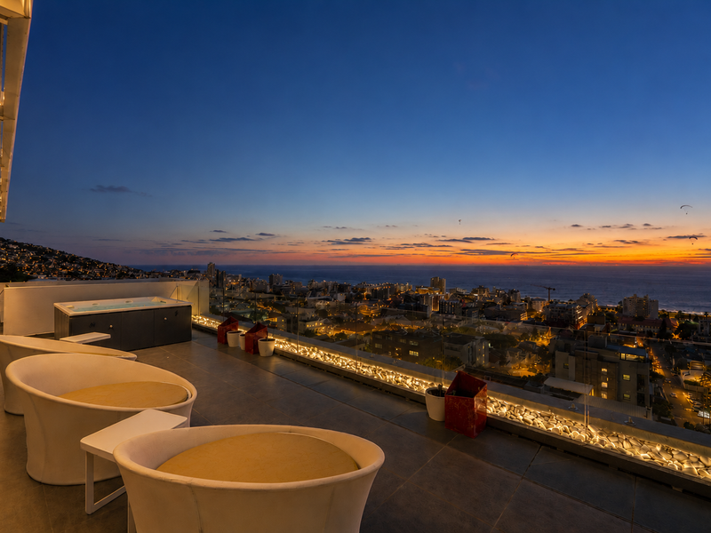Spacious penthouse balcony featuring a built-in jacuzzi, glass balustrades, and panoramic sunset ocean and city views.