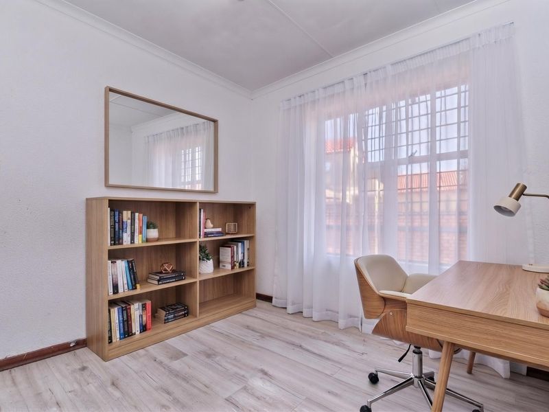A bright home office featuring light-toned laminate flooring, neutral walls, and a large window with security bars, providing a functional workspace.