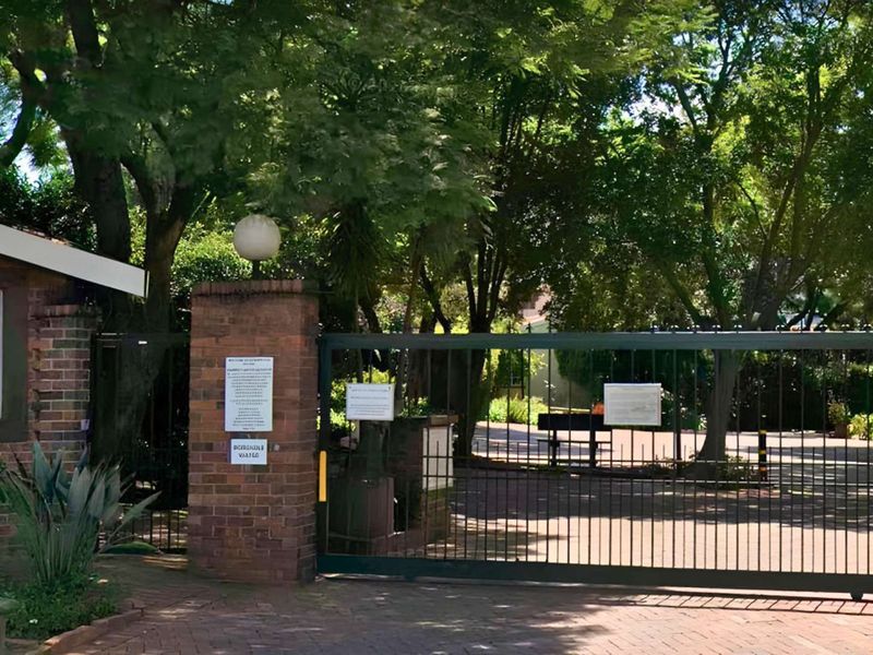 A secure gated entrance to the apartment complex featuring brick pillars, metal security gates, and mature trees providing shade.