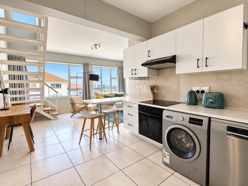 Modern open-plan kitchen and living area featuring sleek white cabinetry, integrated appliances, tiled flooring, and a staircase leading to an upper level.