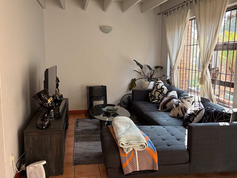 A cozy living room featuring exposed beam ceilings, tiled flooring, and large windows with security bars, providing natural light to the space.