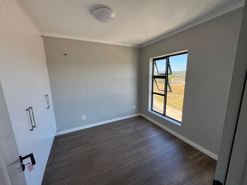 A clean, empty bedroom featuring modern laminate flooring, built-in cupboards, and a large window with views of the surrounding area.