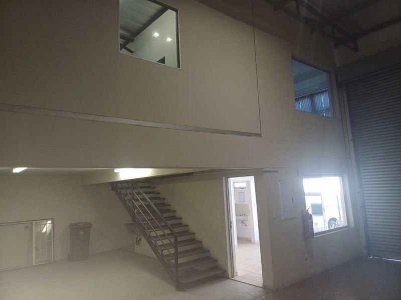 Industrial warehouse space featuring a mezzanine level, metal staircase, roller shutter door, and internal office windows, providing versatile storage or workspace potential.