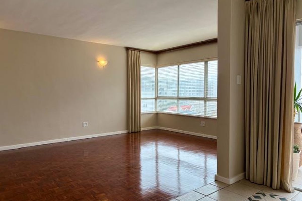 Spacious living area featuring polished parquet flooring, large corner windows providing natural light, and neutral wall finishes.