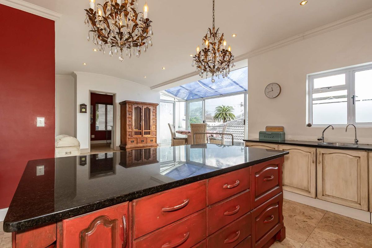 Spacious kitchen featuring a large central island with granite countertops, tiled flooring, and decorative chandeliers, opening into a sunlit conservatory area.