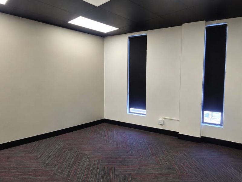 A clean, empty office space featuring modern black ceiling panels, patterned carpet tiles, and tall windows with fitted black roller blinds.