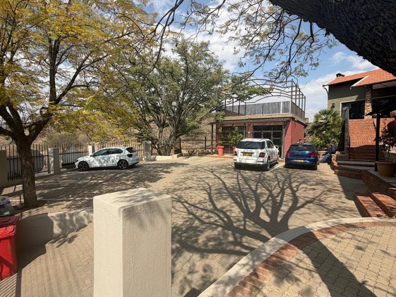 A spacious paved driveway and parking area featuring mature trees, perimeter fencing, and a retaining wall structure.