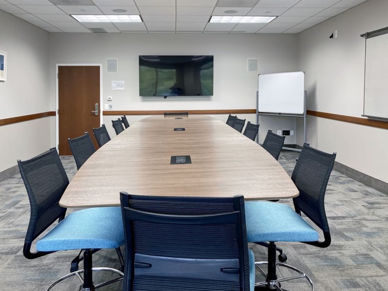 A professional office boardroom featuring a large central conference table, suspended ceiling with recessed lighting, and integrated wall-mounted display screen.