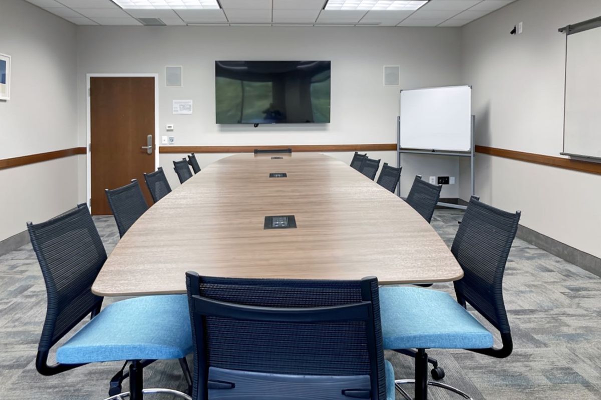 A professional office boardroom featuring a large central conference table, suspended ceiling with recessed lighting, and integrated wall-mounted display screen.