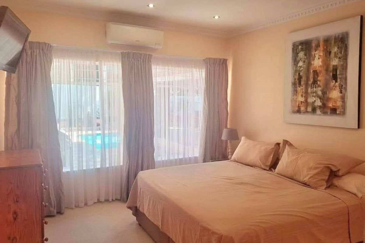 A comfortable bedroom featuring carpeted flooring, recessed downlighting, and a wall-mounted split-system air conditioner, with large windows overlooking the pool area.