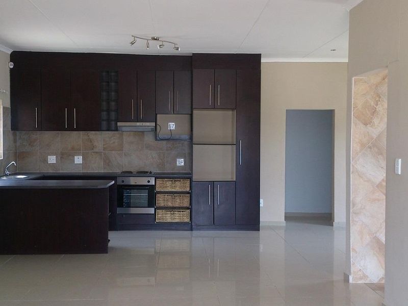 A clean, modern kitchen featuring dark wood cabinetry, built-in oven and hob, tiled backsplash, and neutral floor tiles in an open-plan layout.