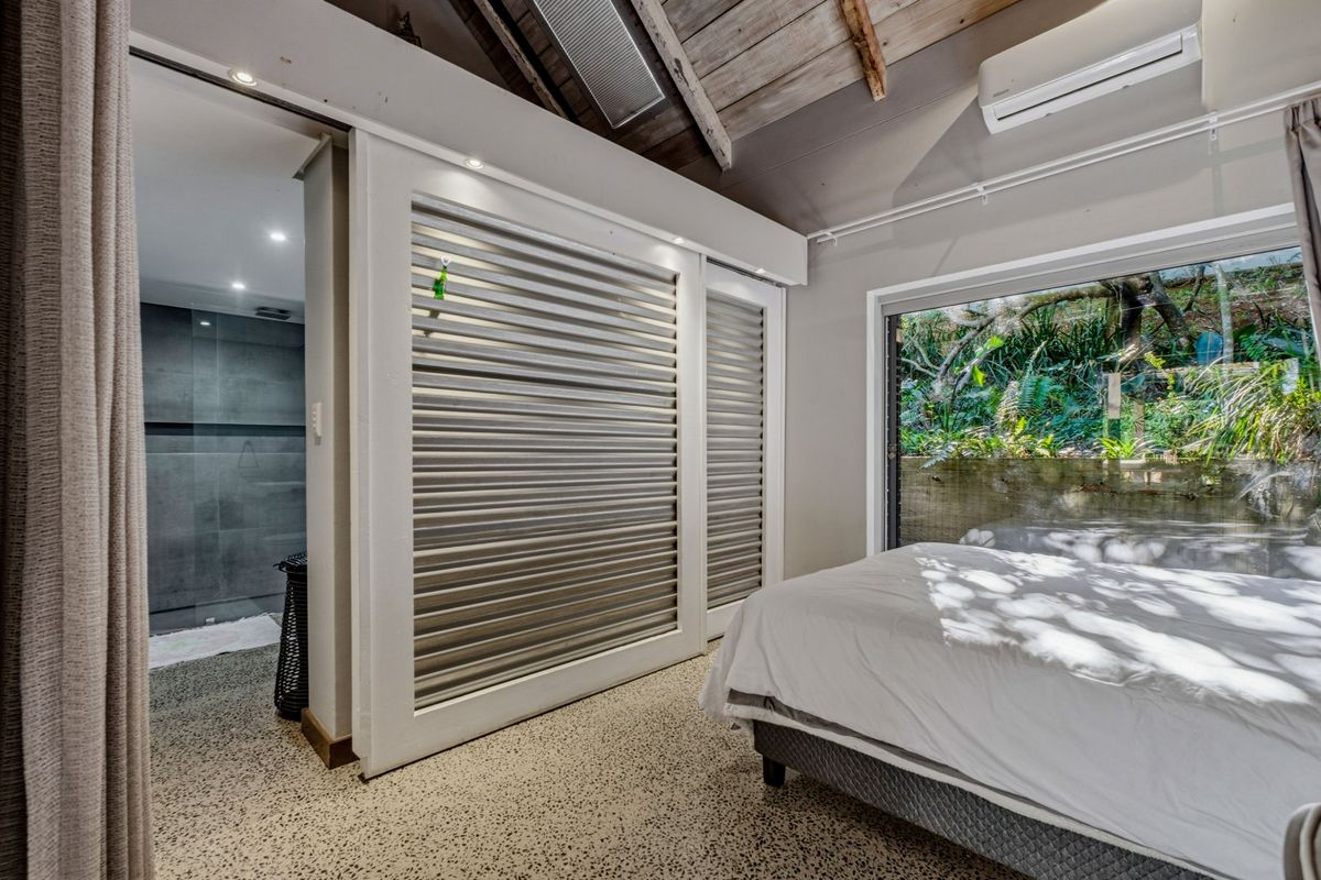 Modern bedroom featuring exposed timber ceiling beams, terrazzo flooring, split-system air conditioning, and sliding louvered shutters leading to an en-suite bathroom.