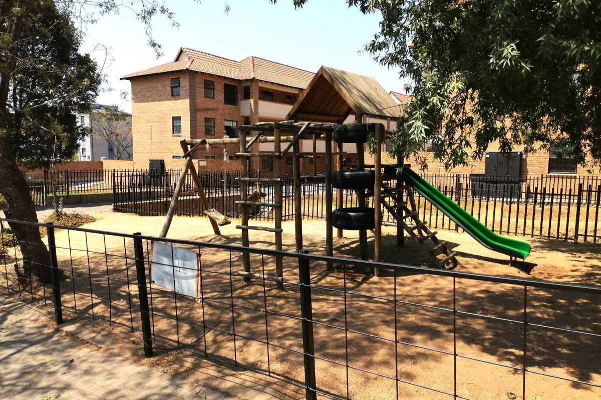 A communal playground area featuring a wooden climbing frame and slide, situated within a fenced complex with brick apartment buildings in the background.