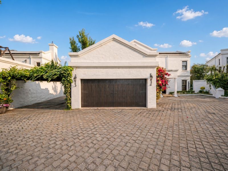 Front view of a well-maintained residence featuring a double garage, cobblestone driveway, and a charming vine-covered pergola.