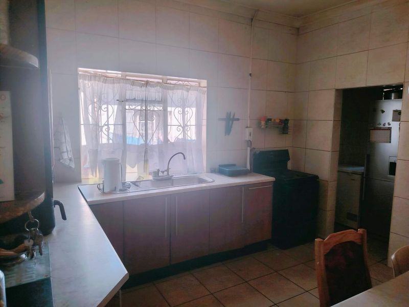 A functional kitchen featuring tiled walls, built-in cabinetry, a stainless steel sink, and a window with security bars, leading into a separate scullery area.