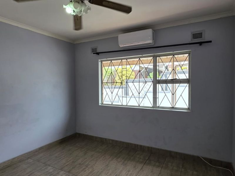 An empty bedroom featuring tiled flooring, a ceiling fan, and a wall-mounted air conditioning unit with security bars on the window.