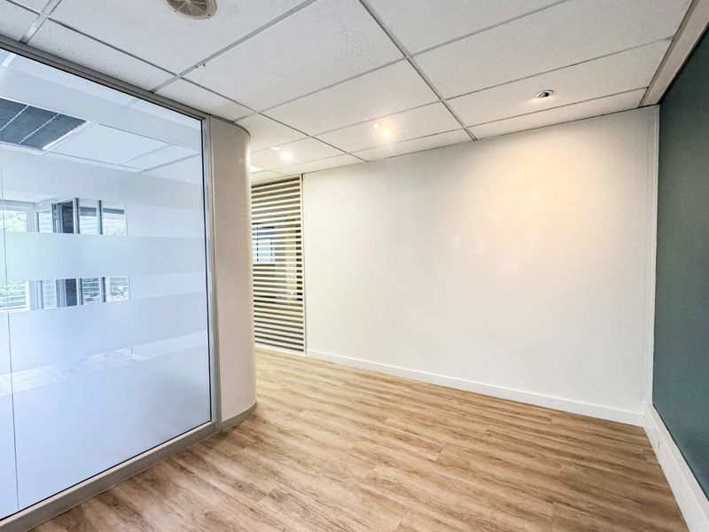 A modern, empty office space featuring laminate flooring, a glass partition wall with privacy film, and a clean suspended ceiling with recessed lighting.