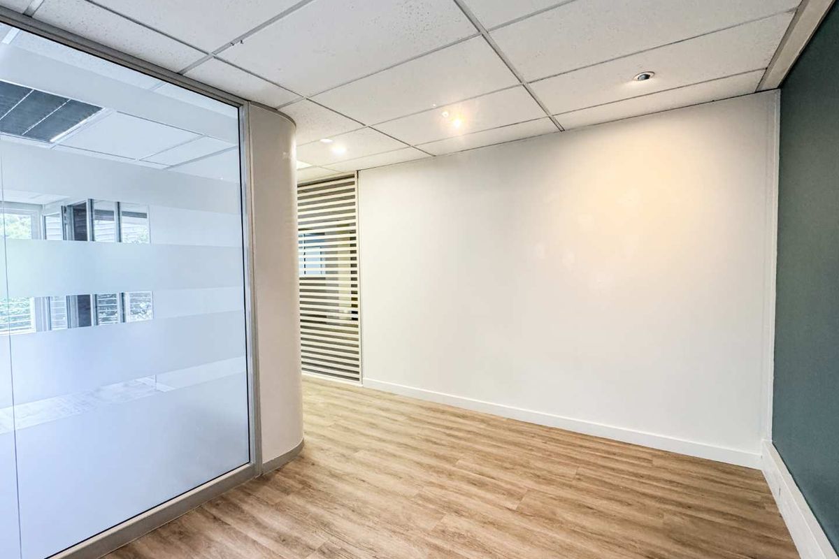 A modern, empty office space featuring laminate flooring, a glass partition wall with privacy film, and a clean suspended ceiling with recessed lighting.