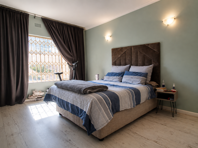 A well-maintained bedroom featuring light-toned laminate flooring, soft wall-mounted lighting, and a large window secured with a metal security gate.