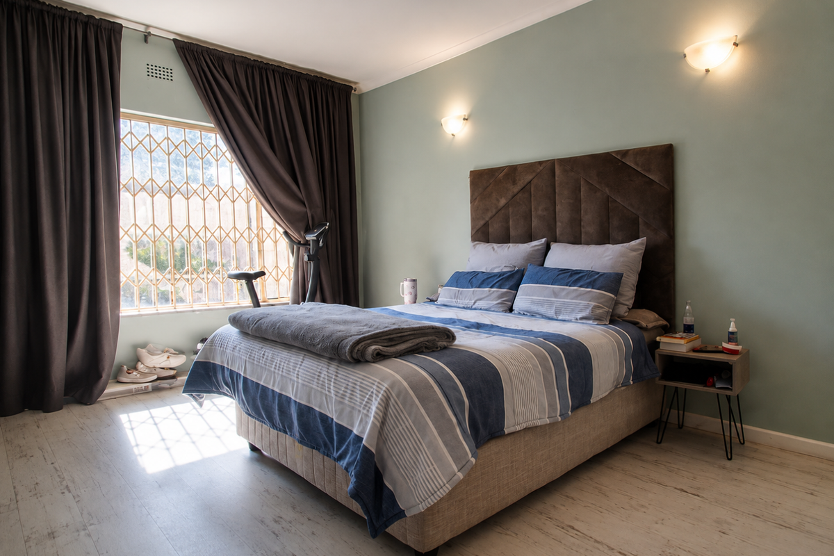 A well-maintained bedroom featuring light-toned laminate flooring, soft wall-mounted lighting, and a large window secured with a metal security gate.