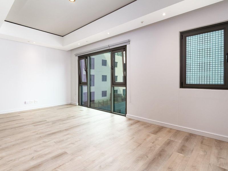A bright, empty living room featuring modern laminate flooring, recessed ceiling lighting, and large windows providing natural light.