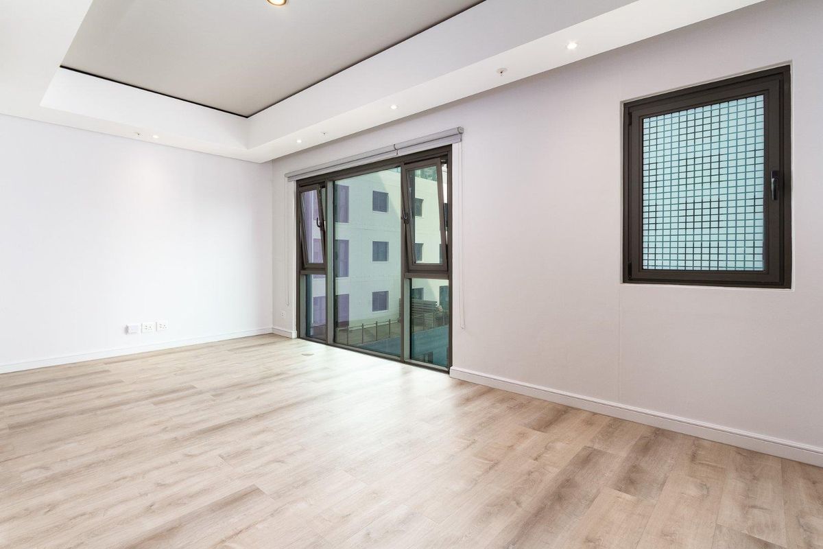 A bright, empty living room featuring modern laminate flooring, recessed ceiling lighting, and large windows providing natural light.