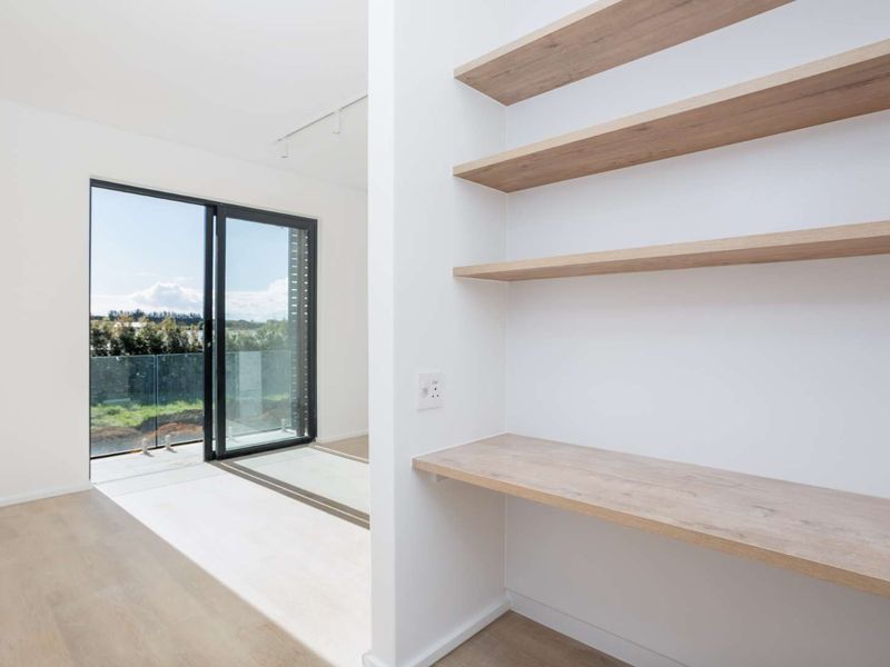 Bright living area featuring built-in timber shelving and desk, with large sliding glass doors opening to a balcony with a glass balustrade.