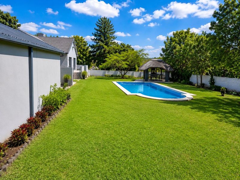 A well-maintained backyard featuring a sparkling swimming pool, a traditional thatched lapa, and a spacious, manicured lawn surrounded by perimeter walls.