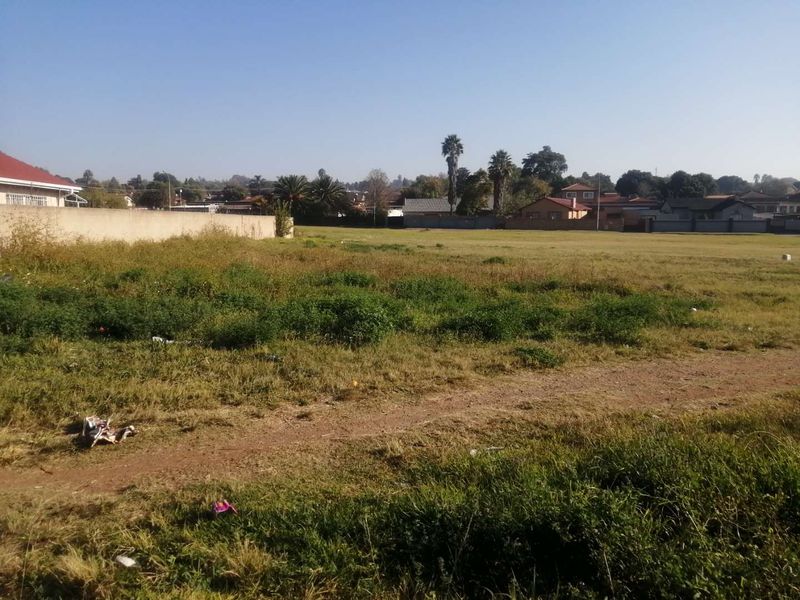 A vacant residential plot featuring open grassland and a perimeter wall, situated in a developed suburban neighborhood.