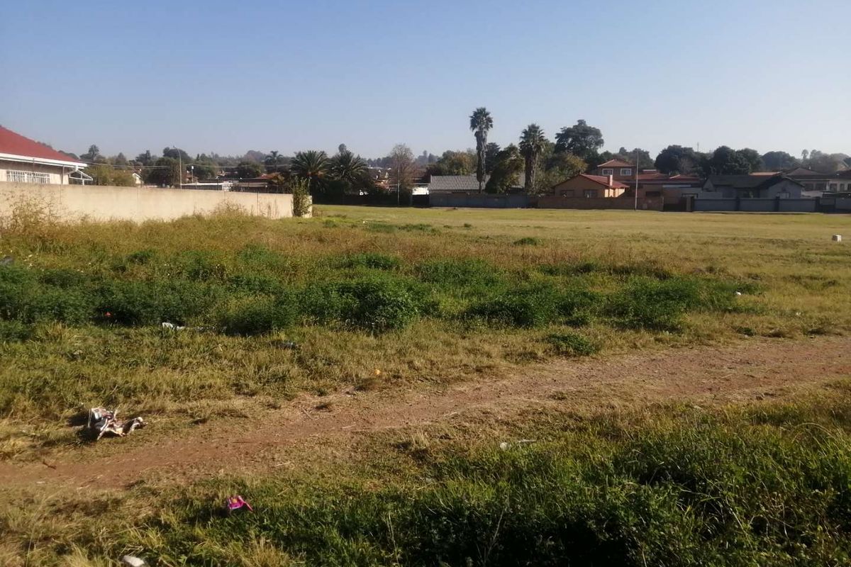 A vacant residential plot featuring open grassland and a perimeter wall, situated in a developed suburban neighborhood.