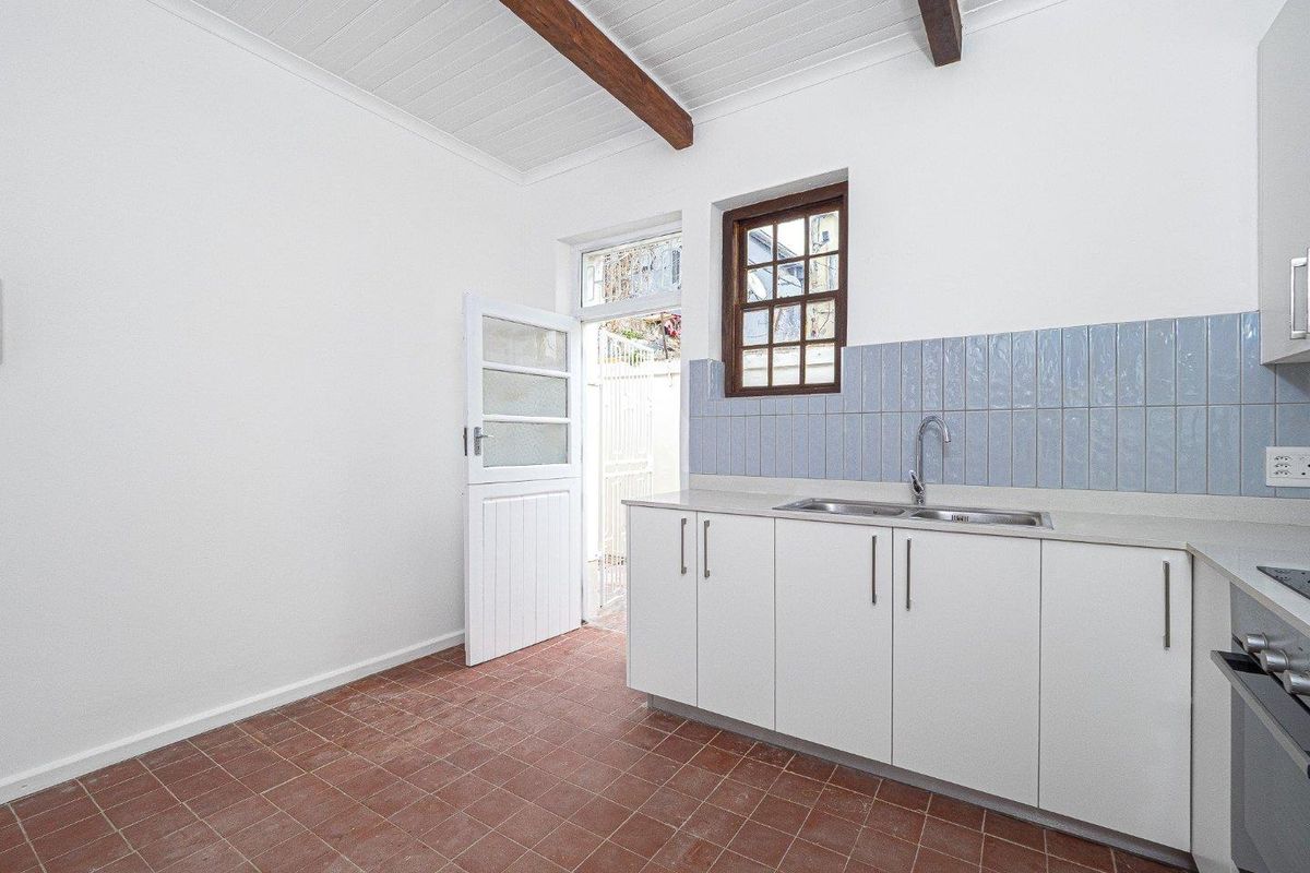 A functional kitchen featuring white cabinetry, a double sink, terracotta flooring, and exposed timber ceiling beams with direct access to an outdoor area.
