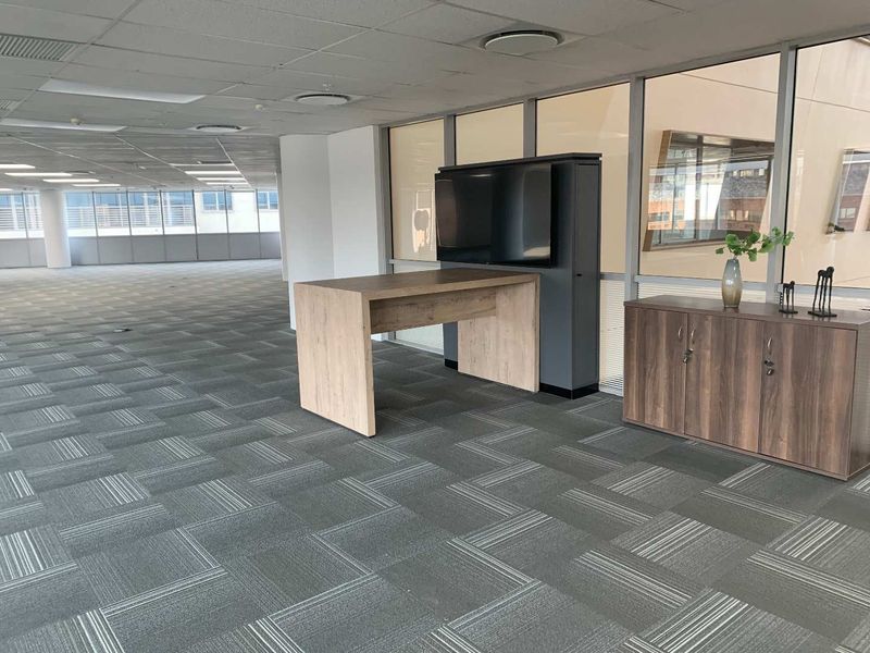 Spacious open-plan office area featuring carpet tiles, suspended ceilings with recessed lighting, and large windows providing ample natural light.
