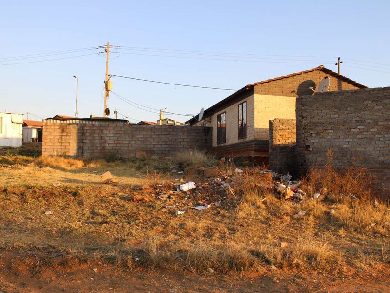 Exterior view of a residential property featuring a brick facade and boundary wall, situated on a plot with significant debris and overgrown vegetation.