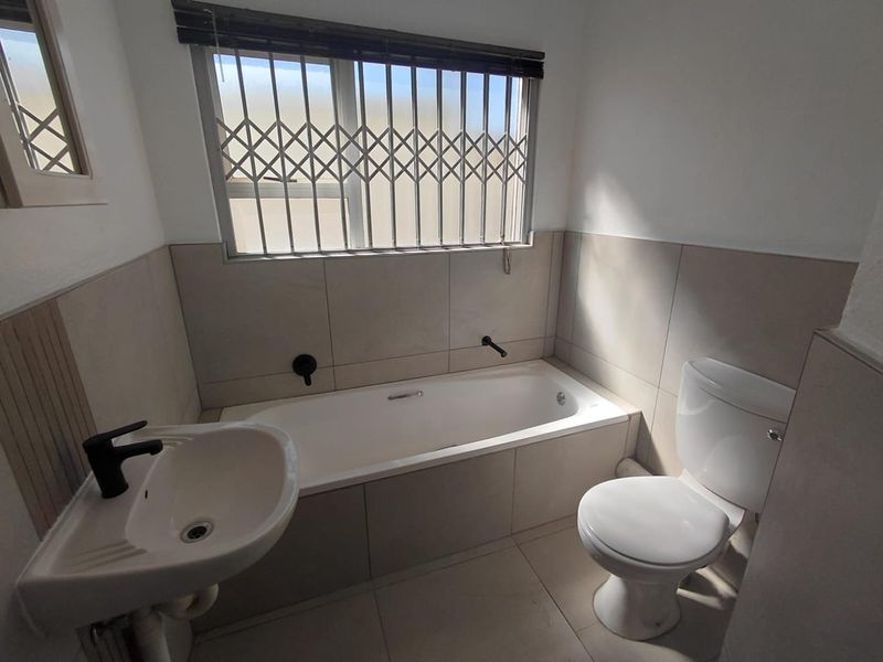 A clean, modern bathroom featuring a built-in bathtub, pedestal basin with black mixer tap, and a standard toilet, finished with neutral wall tiles.