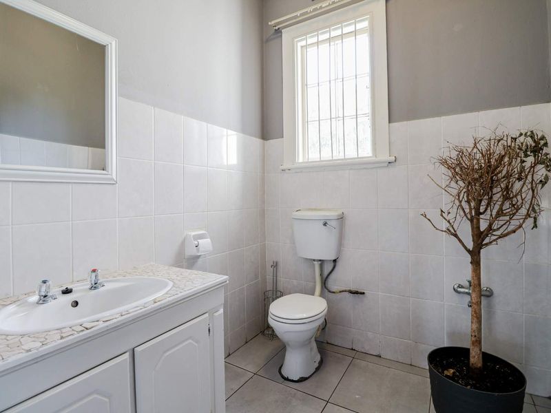 A functional office bathroom featuring a white vanity with integrated basin, toilet, and tiled walls with a security-barred window.