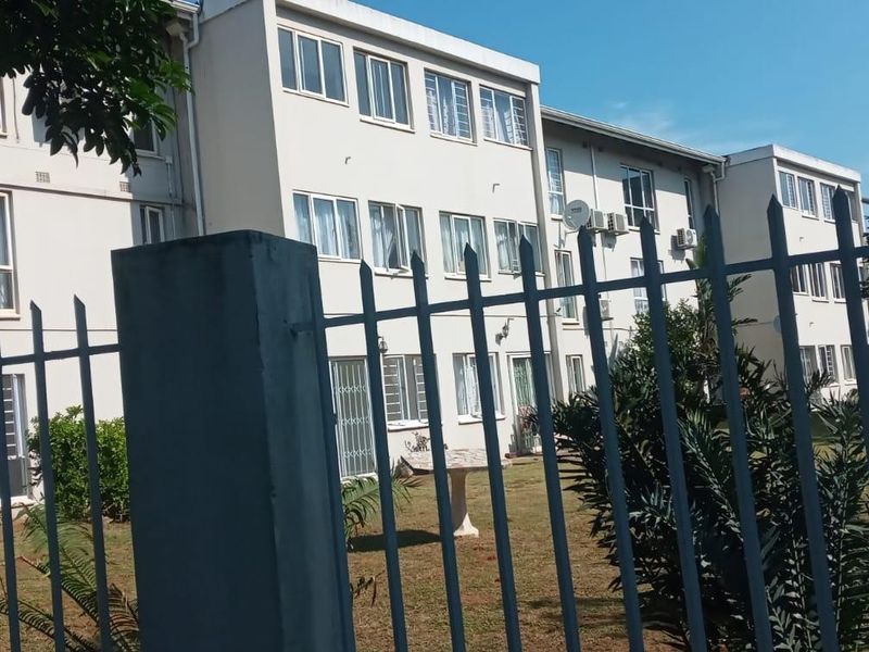 Exterior view of a multi-story apartment building featuring a metal security fence, landscaped grounds, and visible external air conditioning units.