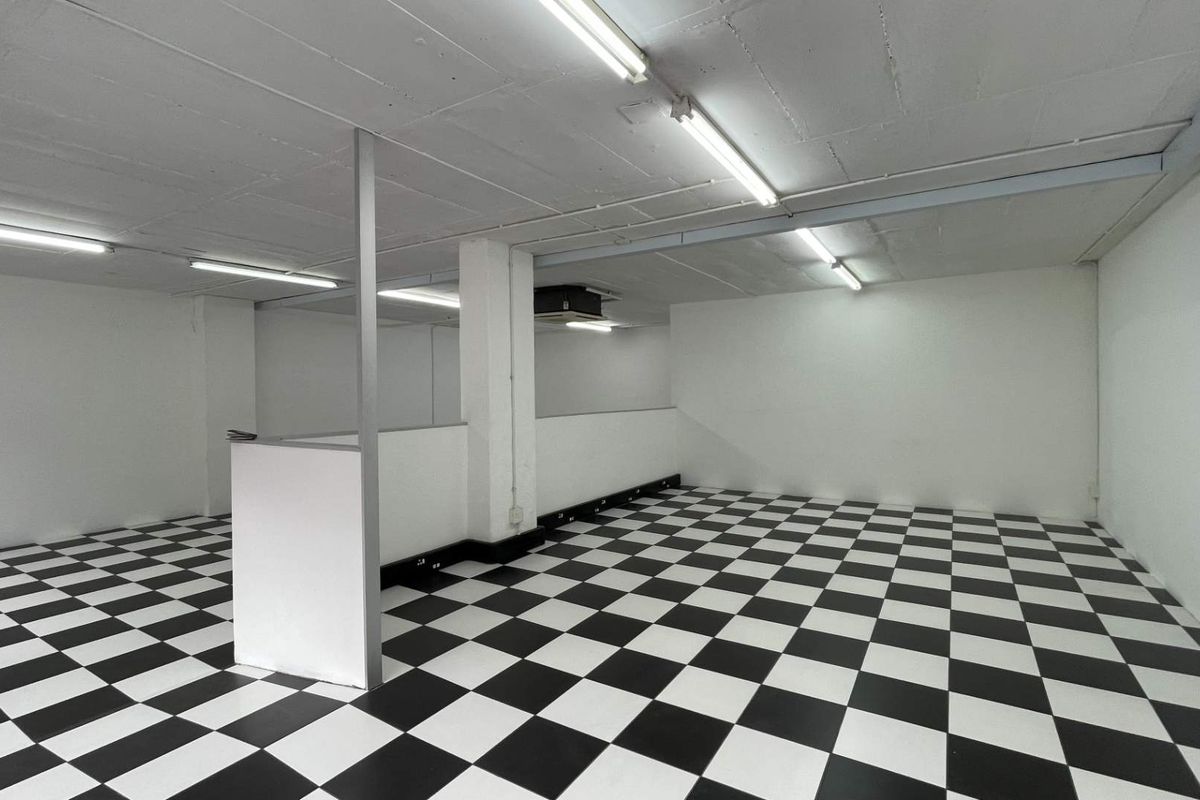 A spacious, empty open-plan retail area featuring high-contrast checkered flooring, exposed concrete ceilings, and functional strip lighting throughout the unit.