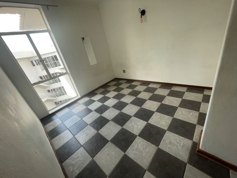 An empty bedroom featuring a large window, checkered tile flooring, and a wall-mounted light fixture, offering a blank canvas for personalization.