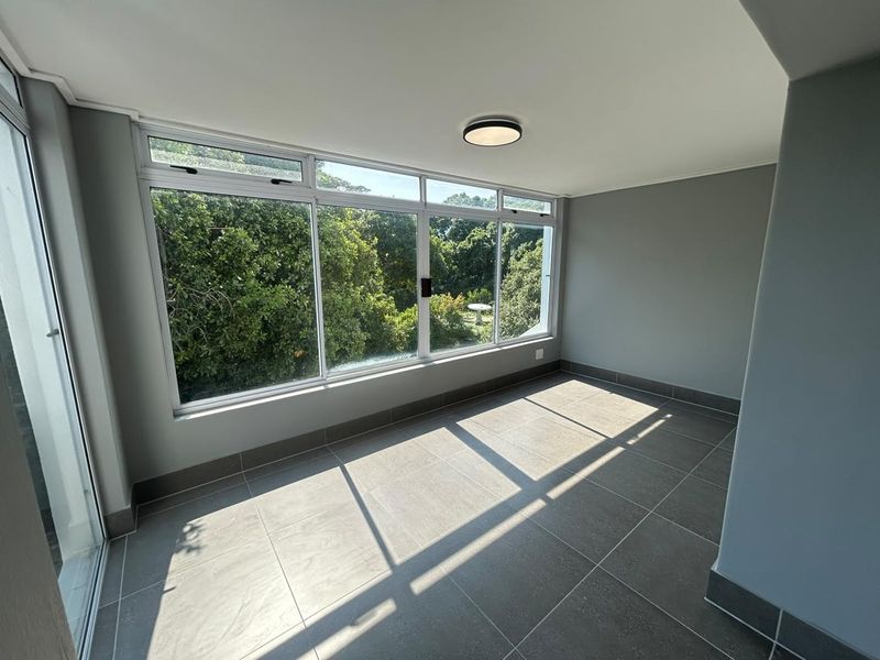 A bright, empty enclosed balcony or sunroom featuring large windows with garden views and modern grey floor tiles.